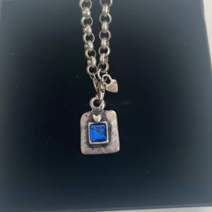 Silverhalsband med blå sten - Snyggt silverhalsband med en fyrkantig blå sten i mitten. Kedjan är robust och har en liten hjärtformad detalj nära hänget. Perfekt för att ge en touch av färg till din outfit. Passar både till vardags och festliga tillfällen.