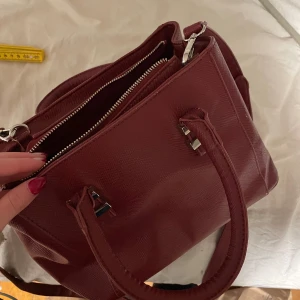 Vinröd väska med dragkedja - Säljer min nyköpta väska. Den är vinröd med silverfärgade detaljer. Väskan har flera fack med dragkedja och handtag.🙌🏻