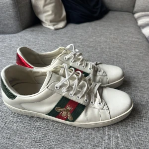 Gucci ace skor  - Säljer ett par vita Gucci sneakers i bra skick. De har gröna och röda ränder på sidan med en broderad bi-detalj. Baksidan av skorna har en röd och grön glansig yta. Perfekta för både vardag och fest! Storlek 41, men passar 42. Tillkommer utan påse och box tyvärr därav priset.
