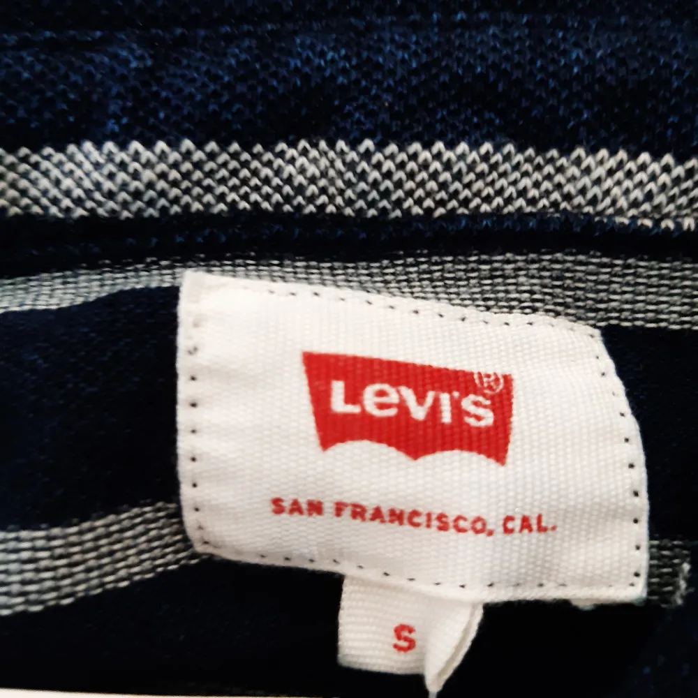 Säljer en snygg blå och vit randig pikétröja från Levi's. Tröjan har en klassisk krage och knappar framtill. Perfekt för en avslappnad stil och passar både till vardags och lite finare tillfällen. Helt ny med taggar kvar!. Skjortor.