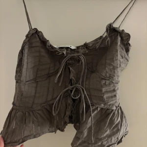 Brun topp från Zara - Säljer en snygg brun topp från Zara i storlek M. Den har tunna axelband och en söt volangdetalj vid halsen. Toppen är croppad och har en knytning framtill som ger en fin passform. Perfekt för sommardagar eller en utekväll!