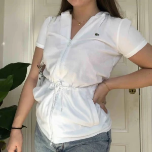 Vit kortärmad hoodie från Lacoste - Säljer en superfräsch vit kortärmad hoodie med luva från Lacoste. Den har dragkedja framtill och en justerbar dragsko i midjan för perfekt passform.  i storlek 42 men passar som en S