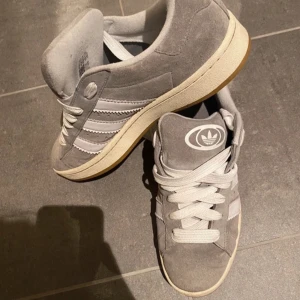 Grå Adidas Campus sneakers - Säljer ett par grå Adidas Campus sneakers i mocka med vita detaljer och snörning. De har en klassisk design med de ikoniska tre ränderna på sidan. Skorna är i bra skick och perfekta för vardagsbruk. Knappast använda, pris kan diskuteras. 