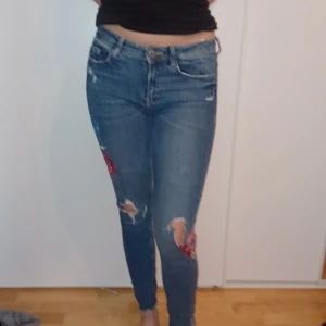 Blå jeans med broderade detaljer - Snygga blå jeans i bra skick med coola broderade blommor på benen. De är tighta och har en låg midja, perfekt för en avslappnad stil. Passar både till vardags och fest!