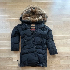 Svart vinterjacka från PJS - Säljer en svart vinterjacka från PJS, Masterpiece Series. Den är i mycket bra skick och har en mysig pälsfodrad huva. Jackan är långärmad och perfekt för kalla vinterdagar. Den har dragkedja och knappar för extra skydd mot kylan. Supervarm och stilren!