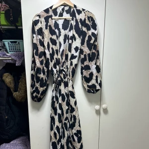 Mönstrad långklänning med knyt - Säljer en superfin långklänning med ett coolt leopardmönster i svart och beige. Klänningen har långa ärmar och ett knyt i midjan som ger en snygg passform. Perfekt för höst och vår, och funkar lika bra till fest som till vardags. Materialet känns mjukt och bekvämt. Hör av dig vid intresse!
