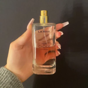 La Rive Her Choice Eau de Parfum - Säljer en elegant och feminin parfym från La Rive, 'Her Choice'. Flaskan är genomskinlig med en gyllene spraypump och rymmer en doft som är både fräsch och blommig. Perfekt för dagligt bruk eller speciella tillfällen. Flaskan är stilren och enkel att ta med i väskan.