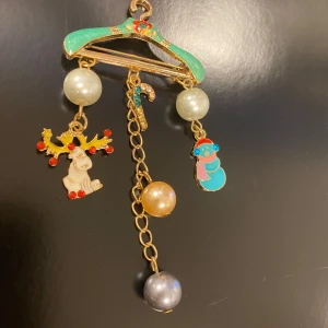 Juligt hängsmycke med ren och snögubbe - Charmigt hängsmycke med julmotiv. Smycket har en grön galge med hängande dekorationer: en ren med röda detaljer, en snögubbe i blått och en polkagris. Pärlor i olika färger ger extra glans. Perfekt för att sprida julstämning!