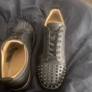 Svarta sneakers med nitar och röd sula - Säljer ett par svarta sneakers i skinn med coola nitar på tån och sidorna. De har en snygg röd sula som verkligen sticker ut. Perfekta för att ge din outfit en edgy touch! Skorna har snörning och är i mycket bra skick.