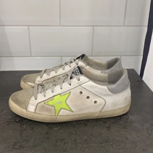 Golden goose sneakers - Säljer ett par Golden Goose sneakers i bra skick. De är vita med en cool neongrön stjärna på sidan och har en vintage-look. Skorna har snörning och är perfekta för en avslappnad stil. Sulan är lite smutsig men det ger dem bara mer karaktär. Perfekta för både vardag och fest!
