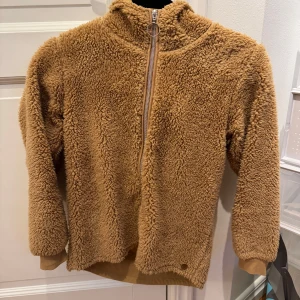 Beige teddyjacka med dragkedja - Mysig beige teddyjacka i storlek 146/152. Perfekt för höst och vinter med sin fluffiga och varma design. Jackan har en praktisk dragkedja framtill och långa ärmar. Den är i mycket bra skick och redo för nya äventyr!