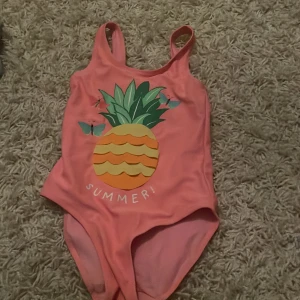 Rosa baddräkt med ananastryck från H&M - Söt rosa baddräkt från H&M med ett färgglatt ananastryck och texten 'SUMMER!' på framsidan. Perfekt för sommarens badäventyr! Passar barn i storlek 110/116, vilket motsvarar 4-6 år. Baddräkten har en enkel design med axelband och är gjord i ett bekvämt material.