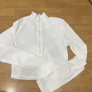 Vit ribbad tröja med dragkedja - Säljer en snygg vit ribbad tröja med dragkedja framtill. Den har en hög krage och långa ärmar, perfekt för en stilren look. Tröjan är croppad och passar bra till både vardag och fest. Skicka ett meddelande vid intresse!