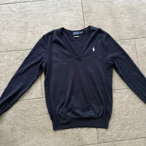 Marinblå tröja från Ralph Lauren - Säljer en stilren marinblå v-ringad tröja från Ralph Lauren. Den har en liten vit logga på bröstet och är i mycket bra skick. Perfekt för både vardag och fest. Passar bra till jeans eller chinos!