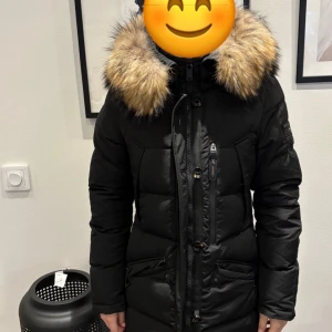 Svart dunjacka med pälsdetaljer - Säljer en snygg svart dunjacka med stor huva och pälsdetaljer. Jackan har långa ärmar och stängs med dragkedja och knappar. Perfekt för kalla vinterdagar. Den är i mycket bra skick och håller dig varm och stilren hela säsongen.