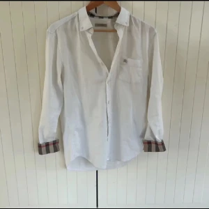 Vit Burberry skjorta - Säljer en stilren vit skjorta från Burberry Brit i storlek S. Skjortan är i 100% bomull och har långa ärmar med klassiska Burberry-rutiga detaljer på insidan av manschetterna och kragen.den har en deffekt där en knapp är sönder mitt på därför det låga priset. Perfekt för både vardag och fest! 🕺