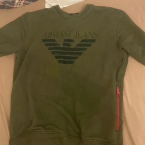 Grön tröja från Armani Jeans - Säljer en snygg grön tröja från Armani Jeans i storlek M. Tröjan har en cool logga på bröstet och en praktisk röd dragkedja på sidan. Perfekt för en avslappnad stil och passar bra till både höst och vår.