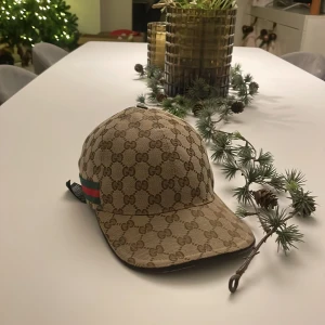 Beige Gucci keps - Snygg beige keps från Gucci med klassiskt monogrammönster. Kepsen har en justerbar rem baktill och en grön-röd randdetalj på sidan. Perfekt för att ge din outfit en lyxig touch. 