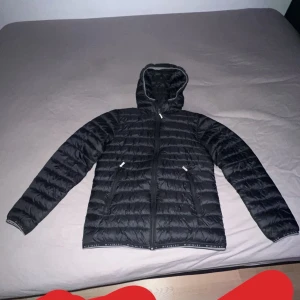 Svart dunjacka från McKinley - Säljer en svart dunjacka från McKinley med huva och dragkedja. Jackan är quiltad och har en stilren design med elastiska kanter. Perfekt för kyligare dagar.