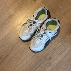 Vita sneakers med guldiga detaljer - Säljer ett par vita sneakers med guldiga detaljer och snörning. Skorna har en svart sula och vit mesh med textdetaljer på sidan. Perfekta för en sportig look.