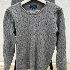 kabelstickad tröja Ralph Lauren - Säljer en stilren grå kabelstickad tröja från Ralph Lauren. Tröjan har en klassisk rund halsringning och är tillverkad i 100% bomull. Perfekt för kyligare dagar. Tröjan är xl i barn storlek så skulle säga att den passar någon med storlek xs.