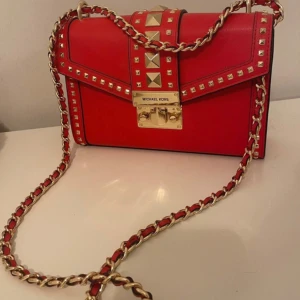 Röd axelväska från Michael Kors - Snygg röd axelväska från Michael Kors med guldfärgade nitar och kedjedetaljer. Väskan har en elegant låsdetalj framtill och är perfekt för att ge en färgklick till din outfit. Insidan är rymlig med flera fack för enkel organisering.