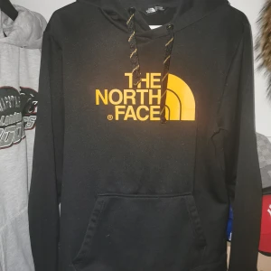 Svart hoodie från The North Face - Säljer en svart hoodie från The North Face med stor gul logga på framsidan. Den har en justerbar huva med snören och en praktisk magficka. Perfekt för kyliga dagar och en stilren look.