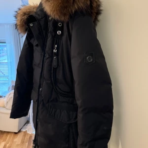 Svart parkajacka från Hollies - Snygg svart parkajacka från Hollies med pälskantad huva. Jackan har dragkedja och knappar framtill samt flera praktiska fickor. Perfekt för kyliga dagar. Nypris på den är 3500kr, sparsamt använd. 