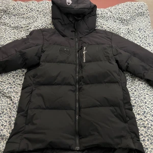 Svart pufferjacka från Sail Racing - Säljer en svart pufferjacka från Sail Racing med huva och dragkedja. Jackan har långa ärmar och logotypdetaljer på bröstet och ärmarna. Perfekt för kyliga dagar.