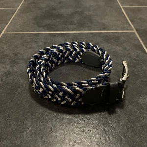 Flätat bälte i blått och vitt - Snyggt flätat bälte i blått och vitt med silverfärgat spänne. Bältet har mörkgråa läderdetaljer vid spännet och ändarna. Perfekt för att ge en stilren touch till din outfit.