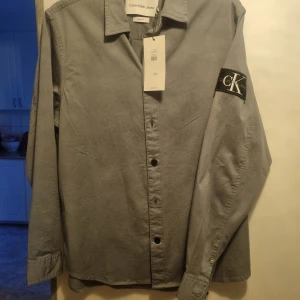 Grå skjorta från Calvin Klein Jeans st M - Snygg grå skjorta från Calvin Klein Jeans med klassisk krage och knappar framtill. Skjortan har en stilren design med ett CK-märke på ärmen. Perfekt för en stilren look.