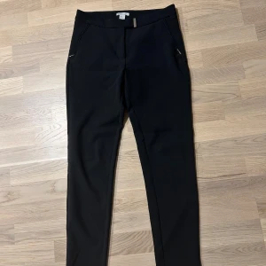 Svarta kostymbyxor med dragkedja - Snygga svarta kostymbyxor med dragkedja vid fickorna. Perfekta för en stilren look. Byxorna har en elegant passform och är ett måste i garderoben.