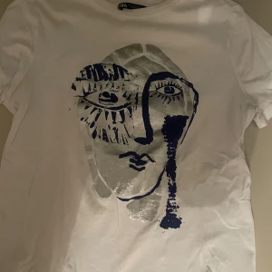 Vit t-shirt med konstnärligt tryck - Snygg vit t-shirt från Zara med ett unikt konstnärligt ansiktstryck i blått och silver. Perfekt för dig som vill ha något annorlunda i garderoben. T-shirten har en normal passform och korta ärmar.