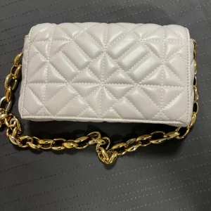 Vit quiltad axelväska med guldkedja - Snygg vit quiltad axelväska med en elegant guldkedja som axelrem. Väskan har en praktisk innerficka med dragkedja och stängs med en tryckknapp. Perfekt för att ge din outfit en lyxig touch.