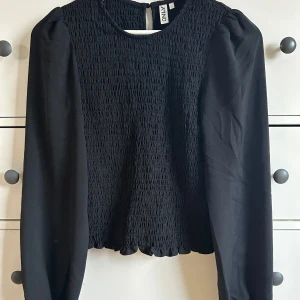 Svart blus från ONLY - Snygg svart blus från ONLY med smockdetaljer och långa ärmar. Perfekt för en stilren look. Blusen har en rund hals och en knapp i nacken för enkel påklädning.