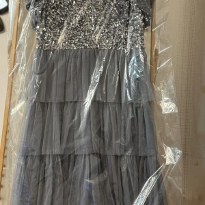 Grå långklänning med paljetter - Elegant grå långklänning med glittrande paljetter upptill och volangdetaljer i kjolen. Klänningen har en v-ringad design och är perfekt för festliga tillfällen.