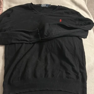 Svart tröja från Polo Ralph Lauren - Säljer en klassisk svart tröja från Polo Ralph Lauren med v-ringning och det ikoniska röda logotypbroderiet på bröstet. Tröjan har långa ärmar och ribbade muddar vid ärmslut och nederkant. Perfekt för en stilren look.