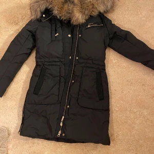 Svart parkajacka med pälsdetaljer - Snygg svart hollies parkajacka med avtagbar huva och pälsdetaljer. Jackan har dragkedja och knappar framtill samt flera praktiska fickor. Perfekt för kyliga dagar. Knappt använd den, säljer pga felstolek