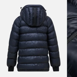 Svart dunjacka från Peak Performance - Snygg och varm svart dunjacka från Peak Performance med huva och dragkedja. Perfekt för kyliga dagar. Jackan har en pufferdesign och långa ärmar. Orginal pris 2100