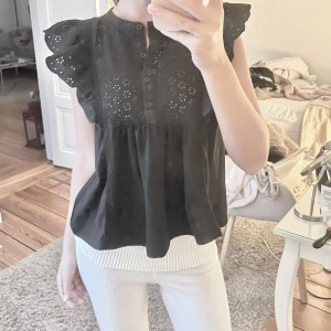 Svart blus med spetsdetaljer - Säljer en söt svart blus med spetsdetaljer och volangärmar. Blusen har knappar framtill och en luftig passform, perfekt för både vardag och festliga tillfällen.