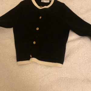 Svart kofta från Zara - Säljer en stilren svart kofta från Zara med kontrasterande vita kanter och guldknappar. Perfekt för att lägga till en touch av elegans till din outfit. Koftan har långa ärmar och en klassisk passform.