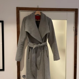 Grå kappa från Mango - Elegant grå kappa från Mango med knytbälte i midjan och bred krage. Perfekt för kyligare dagar med sin stilrena design och långa ärmar.
