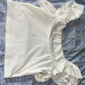 Vit topp med volangdetaljer från Shein - Säljer vit topp från Shein med volangdetaljer vid halsen och axlarna. Toppen har en ribbad struktur.
