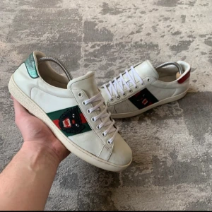 Gucci Ace - Snygga vita sneakers med klassiska gröna och röda ränder på sidan. Skorna har snörning och unika detaljer i grönt och rött på hälen. Perfekta för en stilren look.