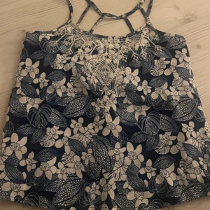 Blommig linne med spetsdetaljer - Snygg blommig linne i blått och vit med vackra spetsdetaljer vid halsen. Perfekt för sommardagar med sina tunna axelband och luftiga passform.