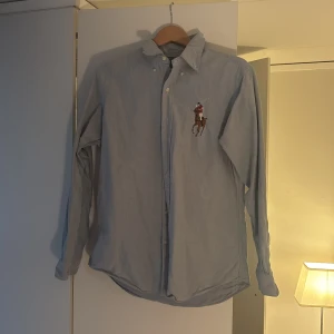 Blå skjorta från Polo Ralph Lauren - Säljer en klassisk blå skjorta från Polo Ralph Lauren med broderad logotyp på bröstet. Skjortan har långa ärmar och knappar framtill. Perfekt för en stilren look.