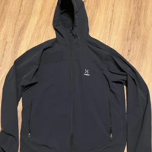 Svart jacka från Haglöfs - Säljer en svart jacka från Haglöfs med huva och dragkedja. Jackan har två fickor med dragkedjor och är perfekt för utomhusaktiviteter. Den är långärmad och har en stilren design.