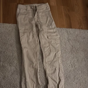 Beige cargobyxor från Kappahl - Säljer ett par beige cargobyxor från Kappahl med flera praktiska fickor och knappstängning. Perfekta för en avslappnad stil med en touch av funktionalitet.