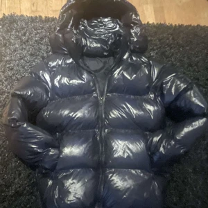 Svart pufferjacka med huva - Säljer en svart pufferjacka med glansig yta och huva. Jackan har dragkedja framtill och är perfekt för kyligare väder. Den är långärmad och har en modern look. Från Everest köpt för ca 1000kr men knappt använd. Modellen är 180cm och väger 67kg.
