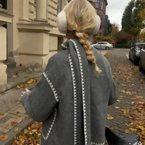 Grå kappa med zigzag-detaljer - Snygg grå kappa med unika vita zigzag-detaljer längs kanterna och fickorna. Kappan har en rak passform och knäpps med knappar framtill. Perfekt för höstpromenader i stan.